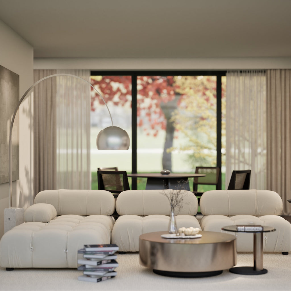 Modern living room with a plush beige sofa, round coffee table, sleek floor lamp, and large windows overlooking autumn trees.