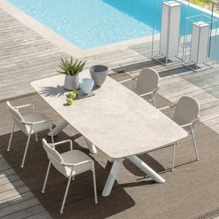 Modern outdoor dining setup featuring a sleek beige stone-top table with white chairs, placed by a poolside on a wooden deck.